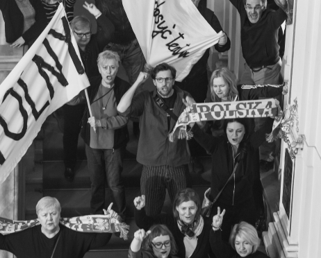 Czarno-białe zdjęcie. Grupa protestujących stoi na schodach w teatrze. Uczestnicy trzymają transparenty i szaliki z napisem „Polska”, część z nich unosi pięści lub wykonuje gest zwycięstwa.